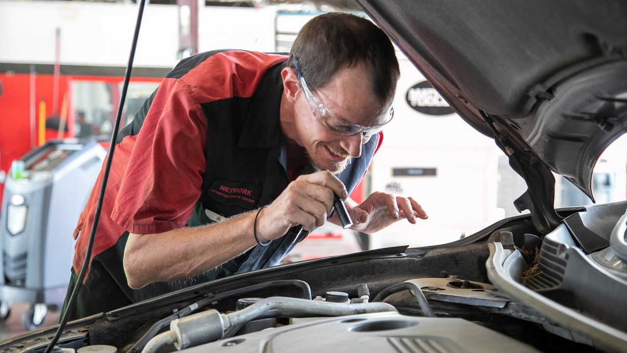 A technician uses a small flashlight to inspect components inside a car's engine bay