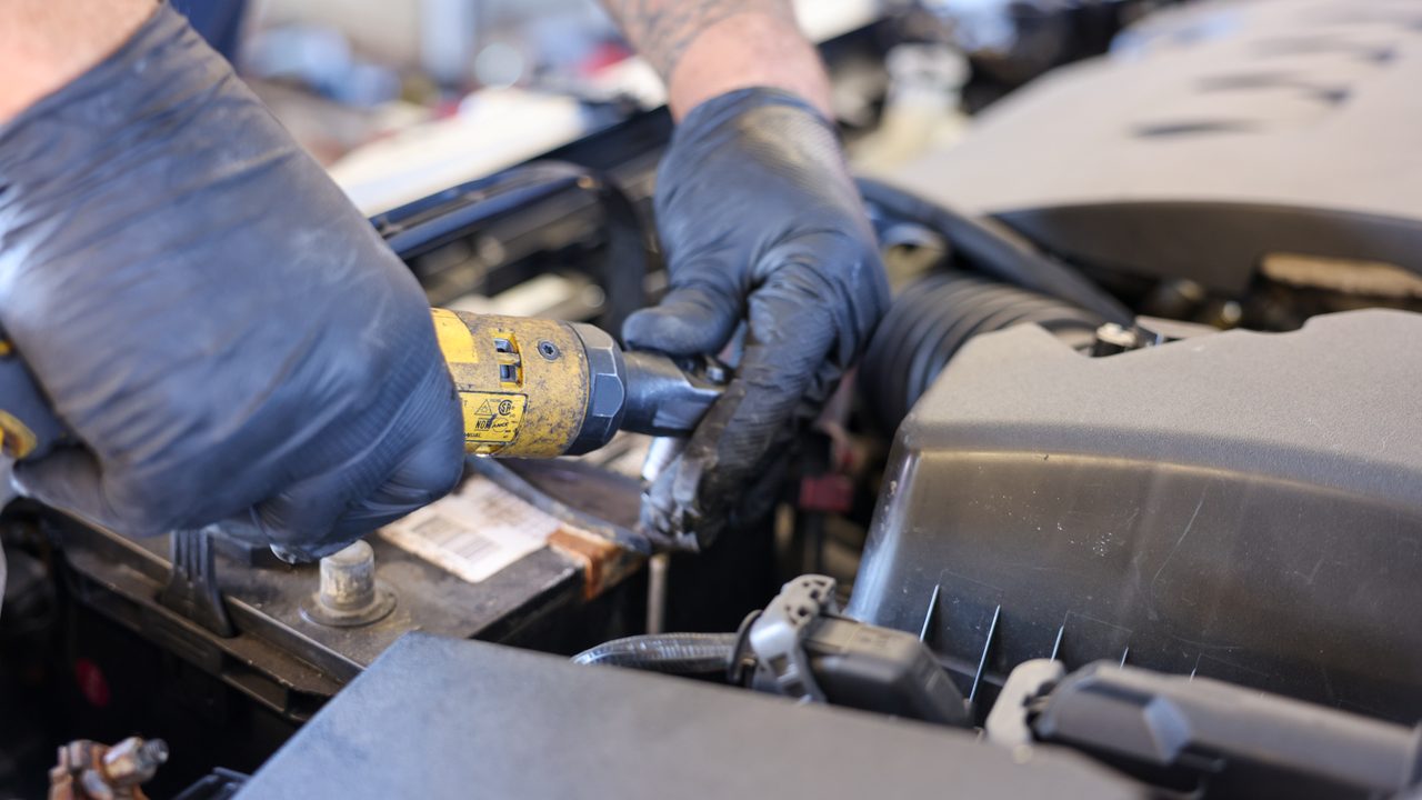 A technician uses a power ratchet to work on components near the battery inside an engine bay
