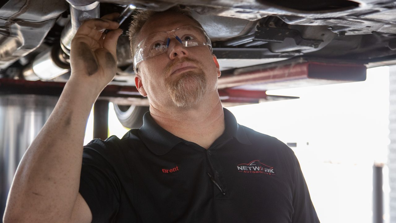 A technician inspects the undercarriage of a raised vehicle using a flashlight in a service bay